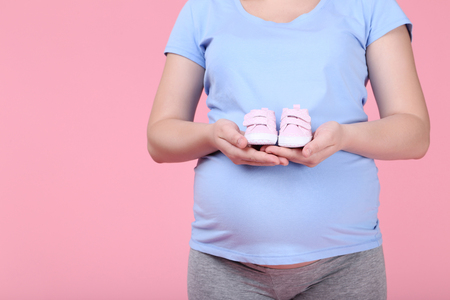 Pregnant woman holding baby shoes on pink backgroundの写真素材