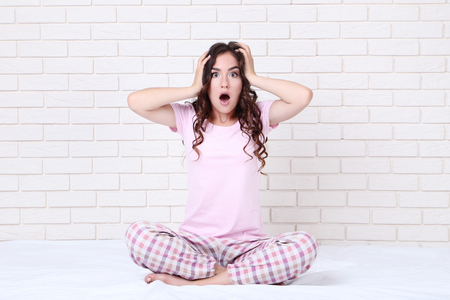 Surprised girl in pajamas sitting on white bedの写真素材