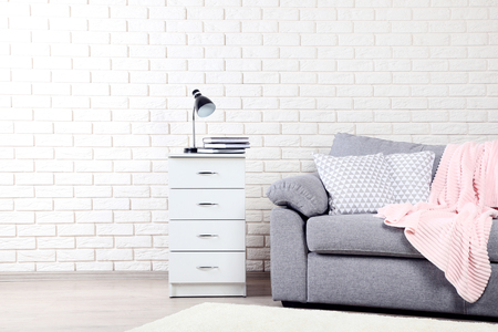 White bedside table near grey sofa with pillows and plaid on brick wall backgroundの写真素材