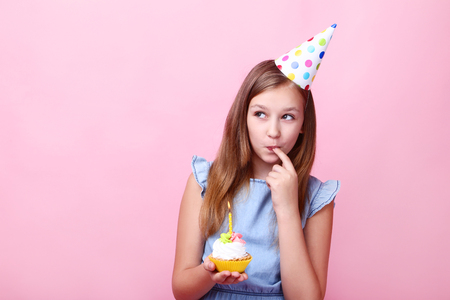 Cute young girl in birthday hat holding cupcake on pink backgroundの写真素材