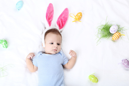 Cute baby with easter eggs and rabbit ears lying on white bedの写真素材