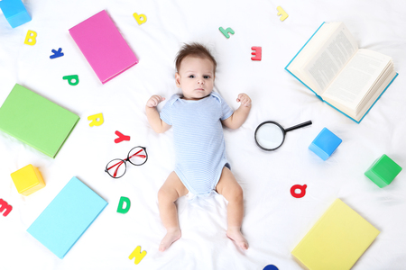 Cute baby with books, glasses, and colored letters lying on white bedの写真素材