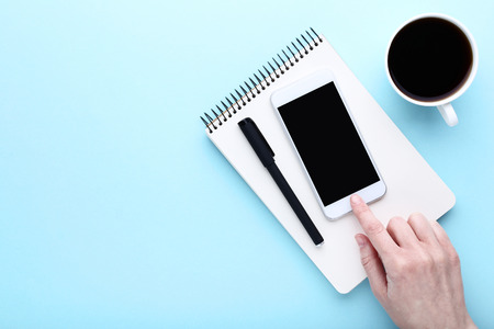 Female finger pressing smartphone button with cup of coffee and notebook on blue backgroundの写真素材