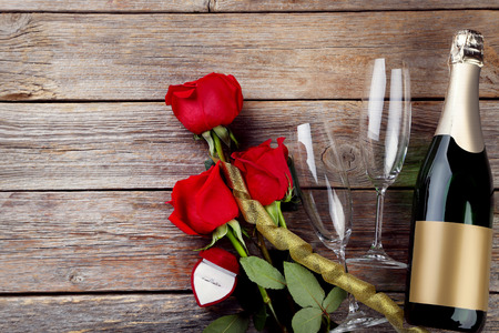 Champagne bottle with glasses, silver ring and bouquet of red roses on wooden tableの写真素材