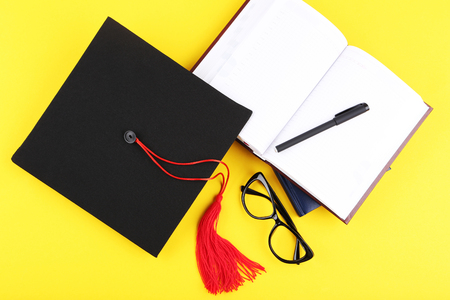 Graduation cap with notepad, pen and glasses on yellow backgroundの写真素材