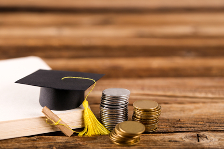 Small graduation cap with notepad and stack of coins on brown wooden tableの写真素材