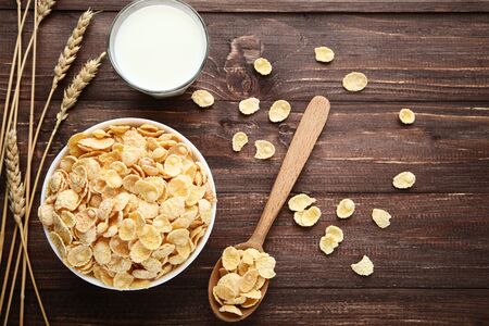 Corn flakes in bowl with wheat ears and glass of milk on wooden table