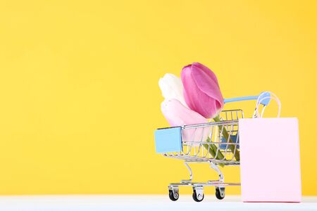 Small shopping cart with tulips and shopping bag on yellow backgroundの写真素材