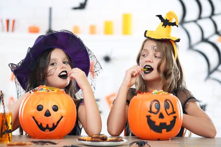 Beautiful two girls in halloween costumes sitting by the table with candies in pumpkin bucketsの写真素材