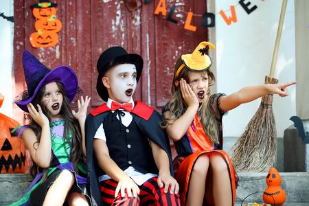 Two surprised girls and boy in costumes sitting on porch with halloween decorationsの写真素材