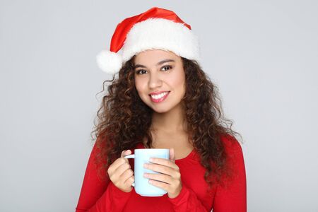 Beautiful american woman wearing santa hat and holding cup of tea on grey backgroundの写真素材