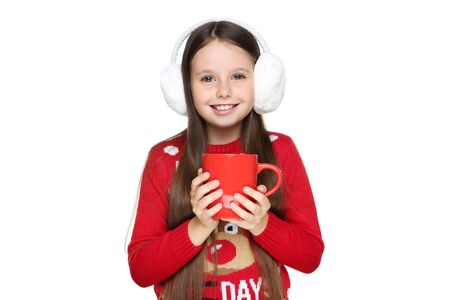 Little girl in christmas sweater and earmuffs holding cup of tea on white backgroundの写真素材