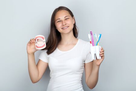 Young girl with dental braces holding teeth model and toothbrushes on grey background