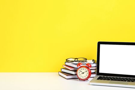Laptop computer with books, glasses and alarm clock on yellow background