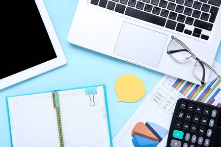Laptop with tablet computer, notepad, calculator and paper charts on blue background