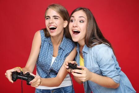 Young happy girlfriends playing computer game with joysticks on red background