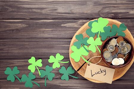 St.Patrick's Day. Paper clover leafs with coins in bowl and word Lucky on wooden table
