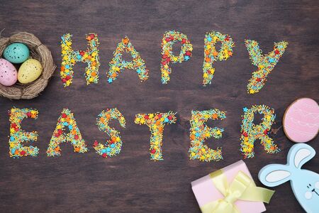 Text Happy Easter with eggs, wooden rabbit and gift box on brown table