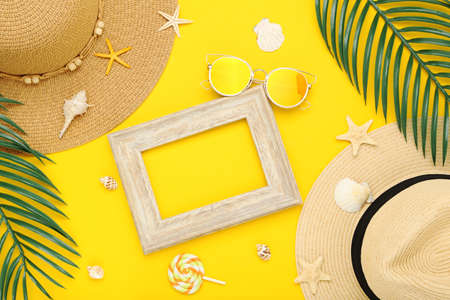 Summer straw hat with palm leafs, sunglasses, seashells and wooden frame on yellow backgroundの写真素材
