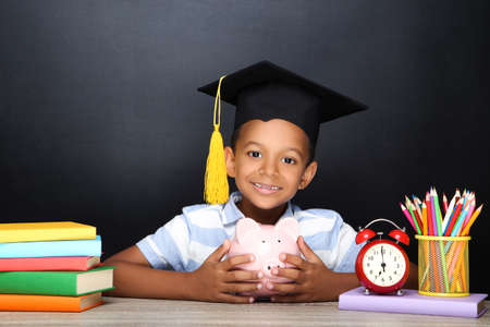Young African American school boy sitting at desk with books, pencils and piggybank on black backgroundの写真素材