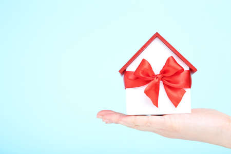 Female hand holding wooden house model with ribbon bow on blue backgroundの写真素材