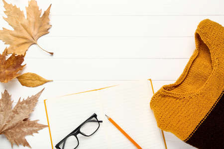 Woolen sweater with eyeglasses, notepad and autumn leafs on white wooden table