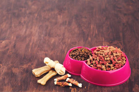 Dry pet food in bowl with bones on brown background