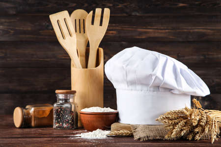 Chef hat with spices in jars, cooking cutlery and bowl with flour on wooden backgroundの写真素材