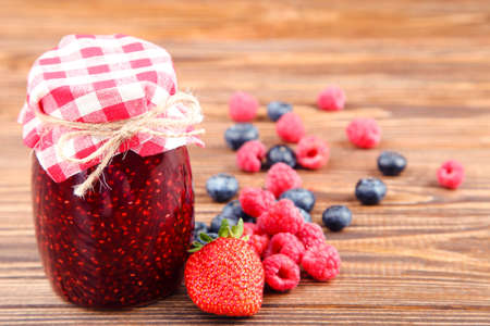 Sweet jam in glass jar with ripe berries on brown wooden background