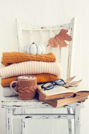 Cup of coffee with knitted sweaters, dry leafs, books and eyeglasses on wooden chairの写真素材