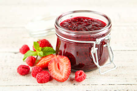 Sweet raspberry jam in glass jar with ripe berries on white wooden table