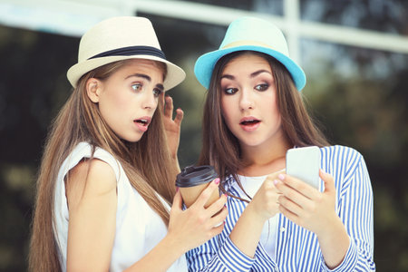 Two young happy girlfriends using mobile phone on the street