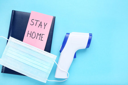 Inscription Stay Home on pink paper, notepad, medicine mask and infrared thermometer on blue...