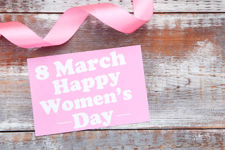 Pink card with text Happy Womens Day and ribbon on wooden background