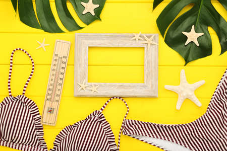Fashion swimsuit with monstera leafs, thermometer, starfishes and photo frame on yellow wooden table