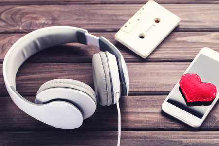 Headphones with smartphone, cassette tape and red heart on wooden background