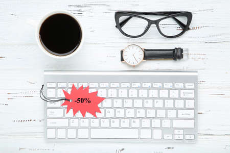 Wristwatch with red tag, eyeglasses, cup of coffee and keyboard on white woden background