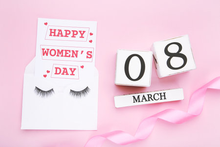 Card with text Happy Women's Day and eyelashes on envelope, wooden cube calendar and ribbon on pink...