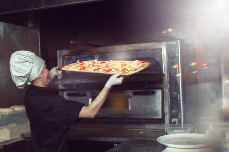 chef baker cook in black uniform putting pizza into the oven with shovel at restaurant kitchen.の写真素材