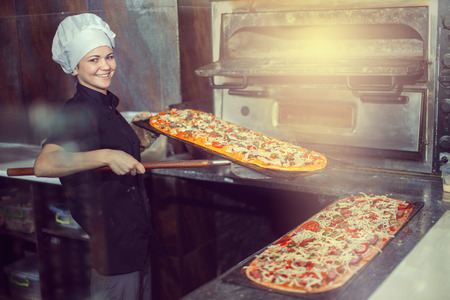 chef baker cook in black uniform putting pizza into the oven with shovel at restaurant kitchen.の写真素材