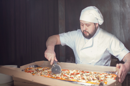 Closeup hand of chef baker in white uniform cutting pizza at kitchen.の写真素材