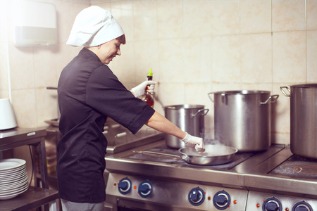 Chef Cooking With Fire In Frying Pan. Professional chef in a commercial kitchen cooking flambe style. Chef frying food in flaming pan on gas hob in commercial kitchen.の写真素材