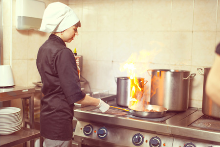Chef Cooking With Fire In Frying Pan. Professional chef in a commercial kitchen cooking flambe style. Chef frying food in flaming pan on gas hob in commercial kitchen.の写真素材