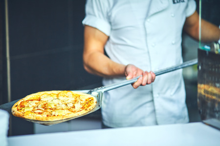 chef baker cook in black uniform putting pizza into the oven with shovel at restaurant kitchen.の写真素材