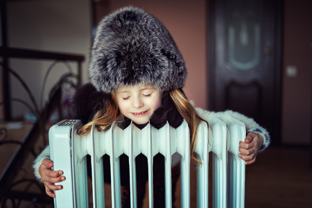 Girl in mittens and hats bask near radiatoの写真素材