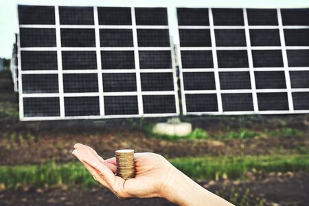 Hand holding money banknote with photovoltaic solar energy panels in background, 