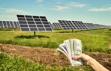 Hand holding money banknote with photovoltaic solar energy panels in background,