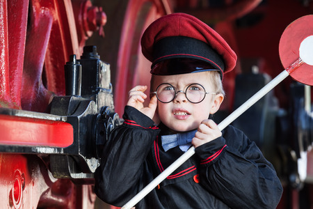 Portrait of a cute little child boy with annoyed facial expression as nostalgic railroad conductor and metalrimmed glasses cap and signaling diskの写真素材