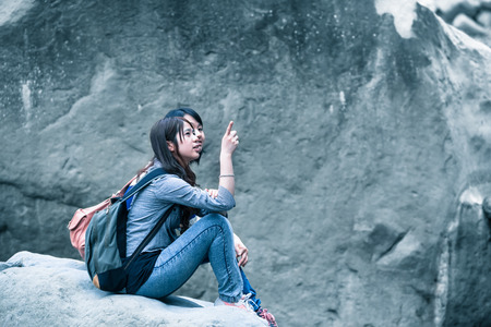 Two young asian women sitting to rest in front of a dark gray wall of rock and talking with gestures by point a finger at something in blue vintage lightの写真素材