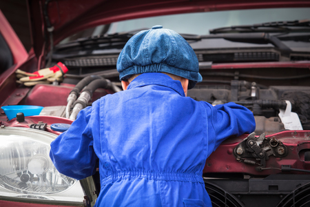 Little boy in blue workwear overall in front of open engine compartment doing some maintenance work at the carの写真素材